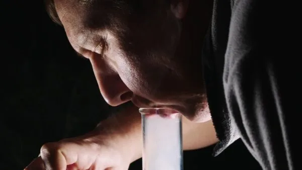Man inhaling white smoke from a test tube with red residue, dark background.