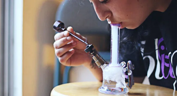 Person inhaling from a glass water pipe, smoke filling the chamber, holding a dab tool.