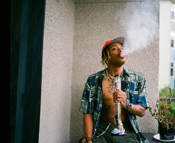 Man with dreadlocks and orange cap smoking a glass bong on a balcony.