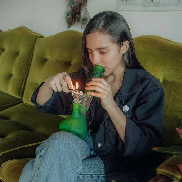 Young woman lighting a green glass bong while sitting on a velvet couch.