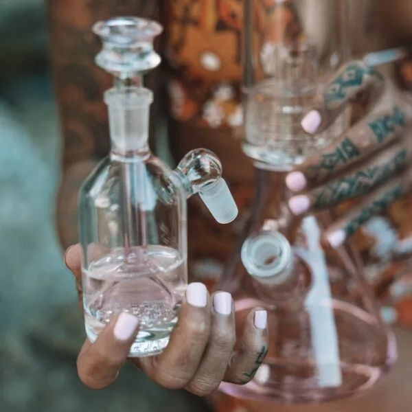 Person holding clear glass ash catcher and pink water pipe with tattooed hands.
