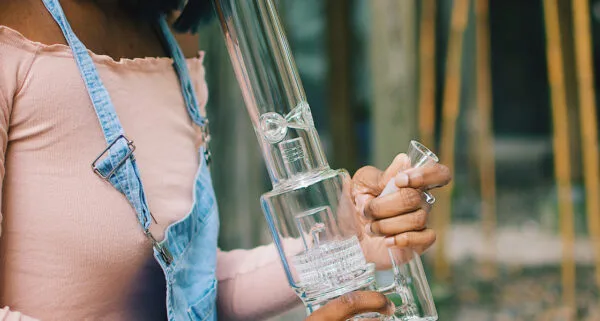 A woman in denim overalls holds a large, clean glass bong with multiple percolators.