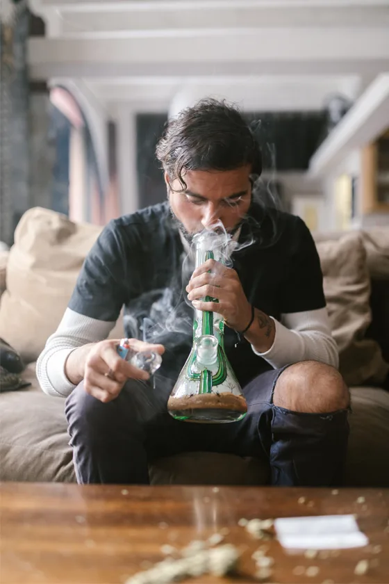 Man smoking cannabis from a green glass bong, relaxing on a couch.