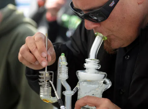 A man with sunglasses using a metal tool to dab cannabis concentrate on a glass rig.