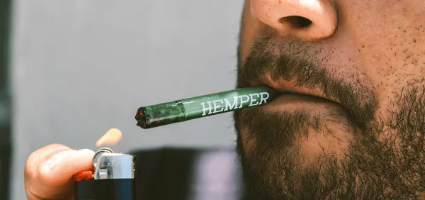 Bearded person lighting a green HEMPER branded pre-roll with a lighter.