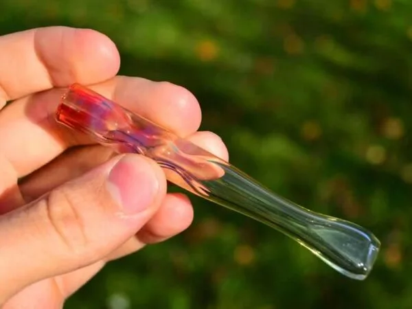 Hand holding a colorful glass smoking pipe with red and green accents outdoors.