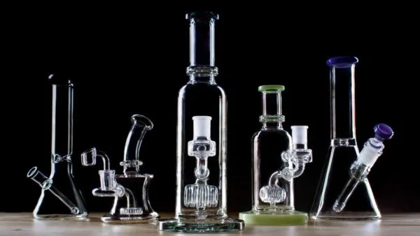 Collection of clear glass bongs, dab rigs, and water pipes on dark background.