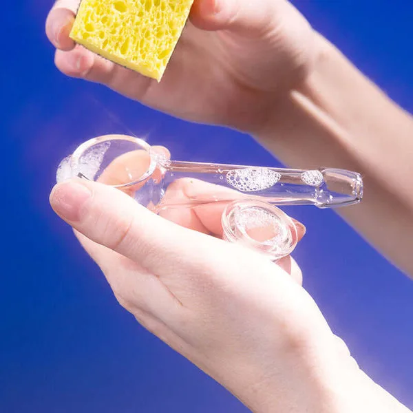 Hands cleaning a clear glass pipe with soapy water and a yellow sponge.