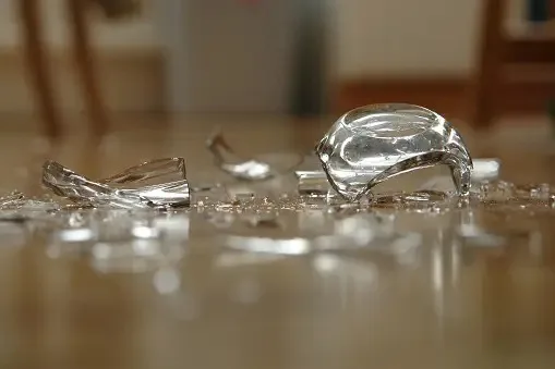 Shattered drinking glass pieces and sharp shards scattered on a wooden floor.