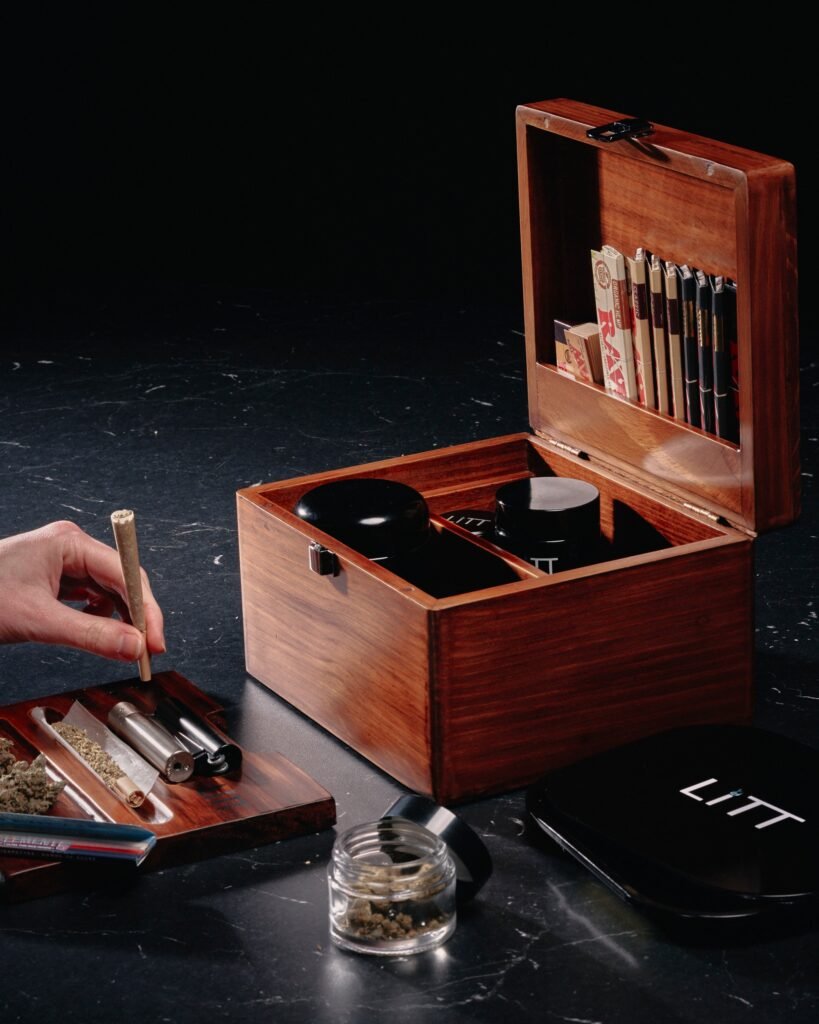 Hand holding pre-roll, wooden stash box with rolling papers, cannabis accessories on dark marble.