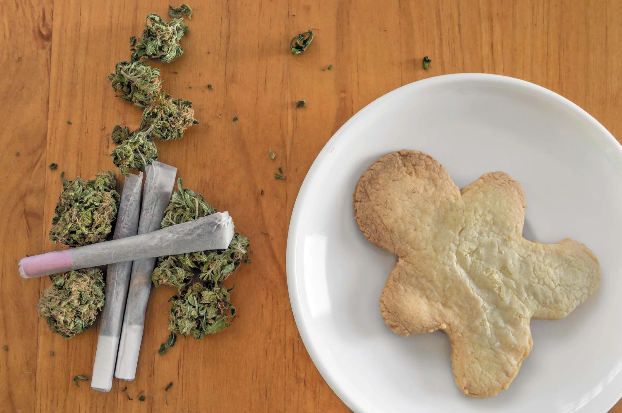 Green cannabis buds, pre-rolled joints, and a cookie on a wooden table.