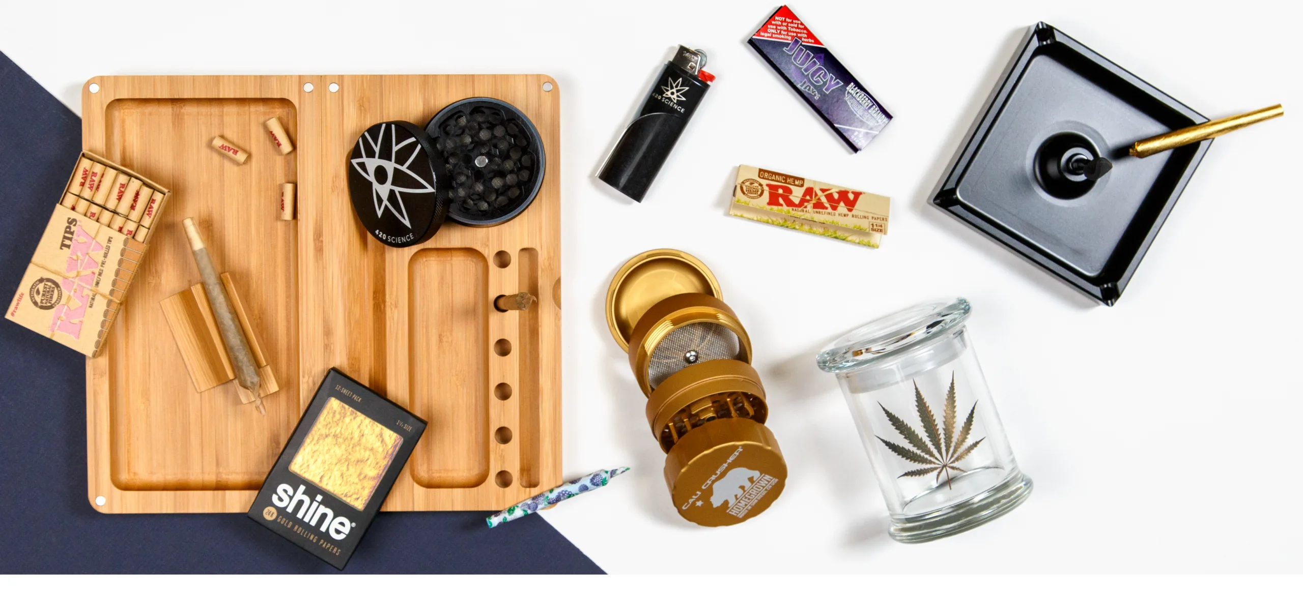 Various cannabis accessories including rolling tray, grinders, papers, lighter, and jar on white background.