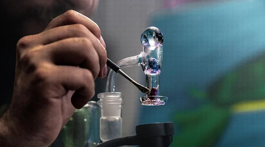 A person's hand using a metal dab tool to load concentrate into a glass rig.