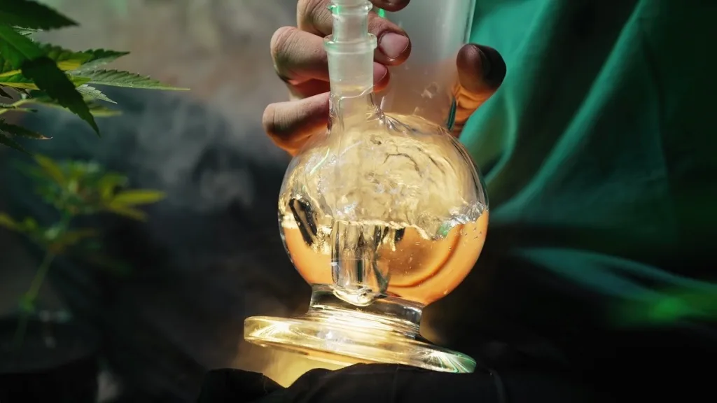 Hand holding bubbling glass bong with cannabis leaves in background.