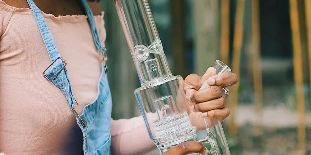 Person in denim overalls holding a clear glass water pipe with intricate percolators and a bowl.