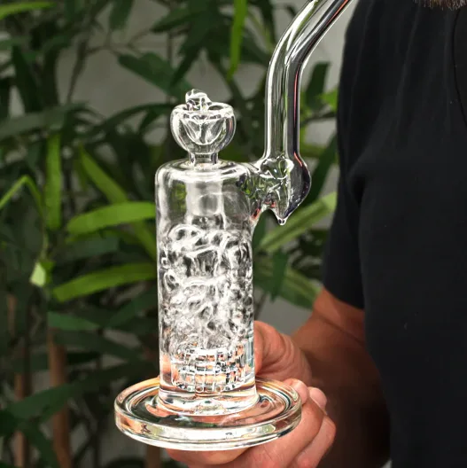 Clear glass water pipe with intricate percolator design, held by hand, green background.