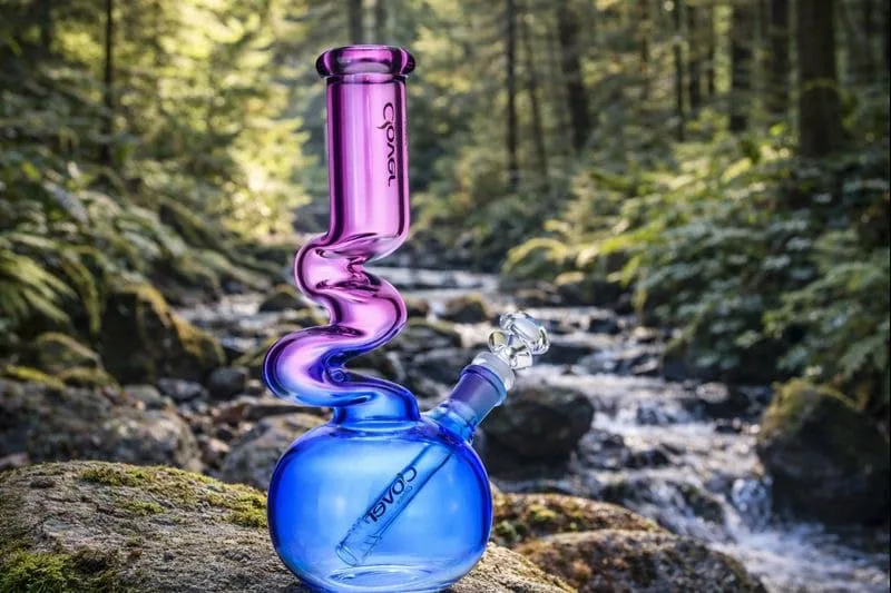 COAST iridescent purple-blue glass water pipe with wavy neck by a forest stream.