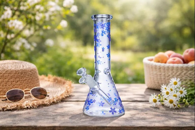 A blue floral glass beaker bong sitting on a wooden table in a garden.