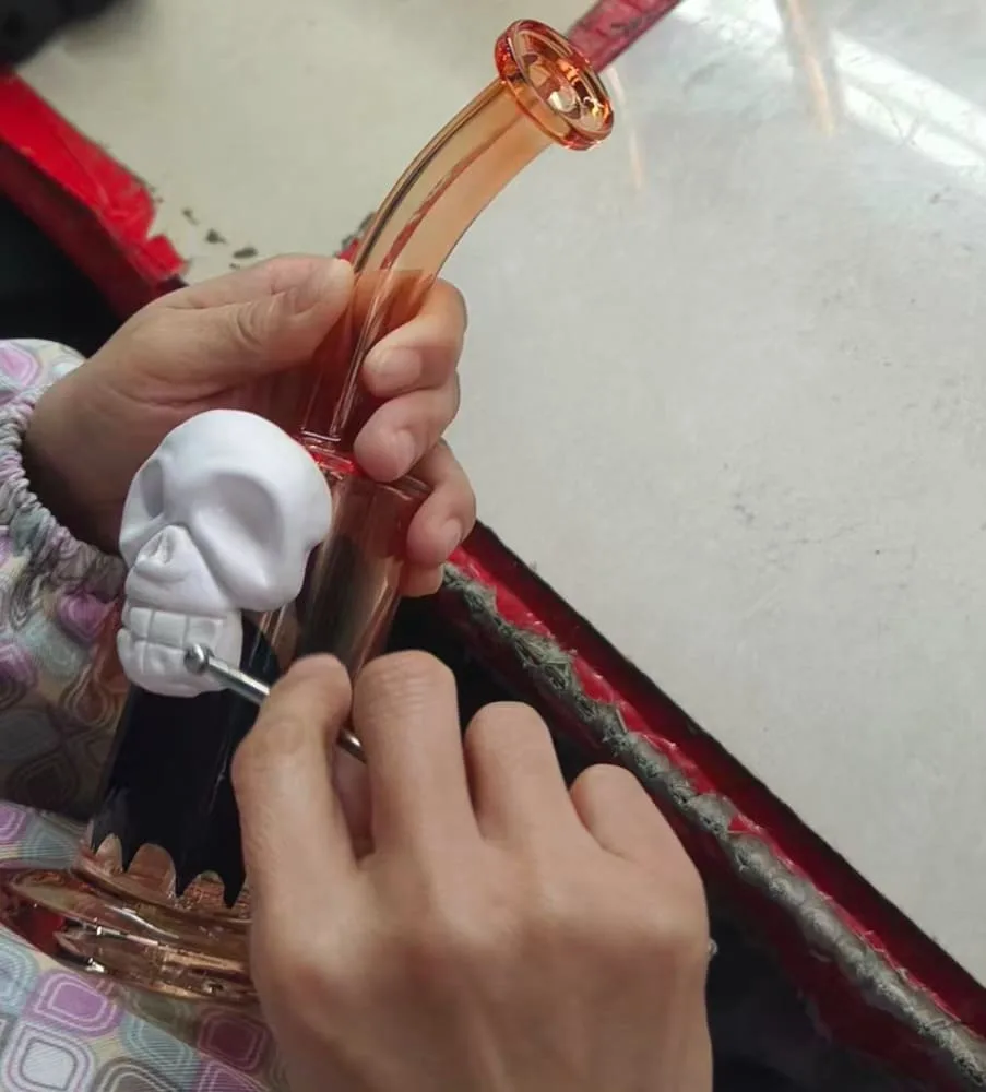 An artisan's hands carefully crafting a custom orange glass bong with a white skull decoration.