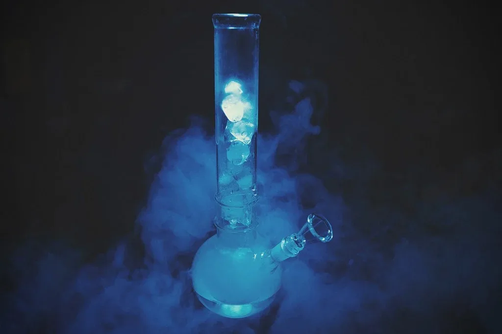 Clear glass bong with ice cubes and blue smoke on a dark background.